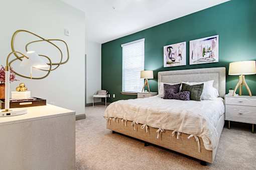 An interior view of a bedroom with a bed, two side tables with lamps on them, a window, and a front end table with books and a lamp at Parkstone at Knightdale apartments for rent in Knightdale, NC.