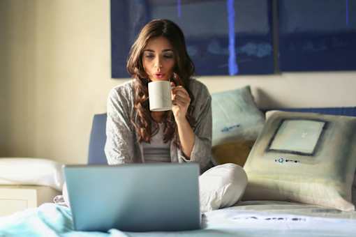 A woman is sitting on a bed with a laptop and a mug in her hand.