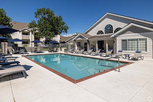 A swimming pool in front of a house with a patio and lounge chairs.