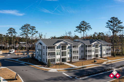A large white building with a grey roof is surrounded by trees and a road.
