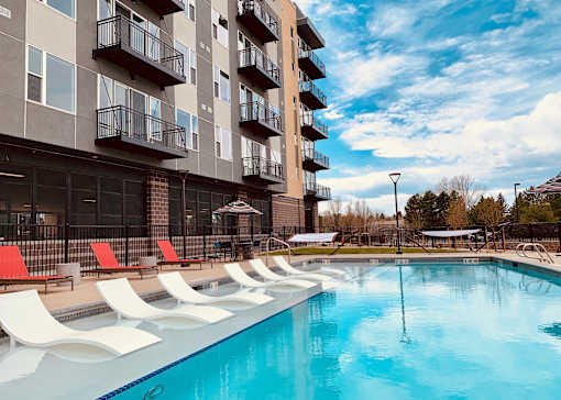 A large swimming pool in front of a building with red lounge chairs.
