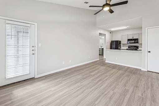 A room with a ceiling fan and light, a door with blinds, and a kitchenette in the background.