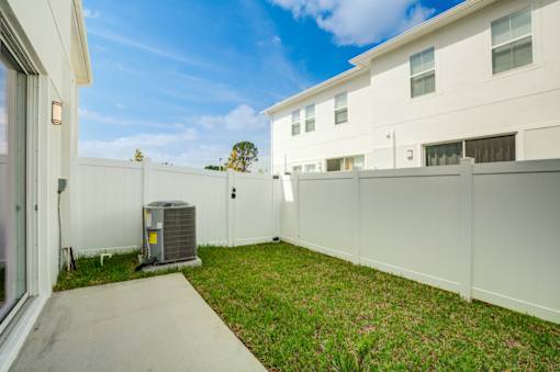 A white fence surrounds a yard with a concrete walkway and a white house in the background.