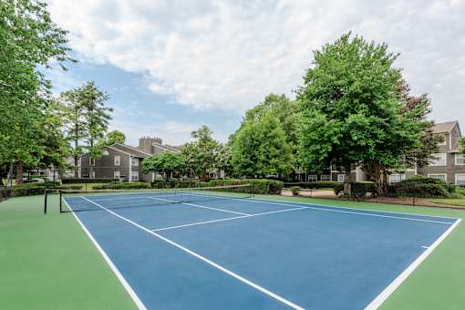 A blue tennis court surrounded by green trees and buildings.