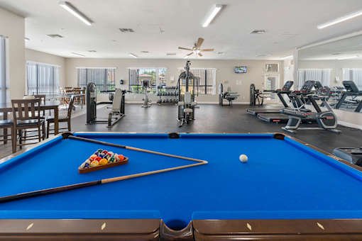 A pool table with a pool ball and cue sticks in the foreground and a gym in the background.