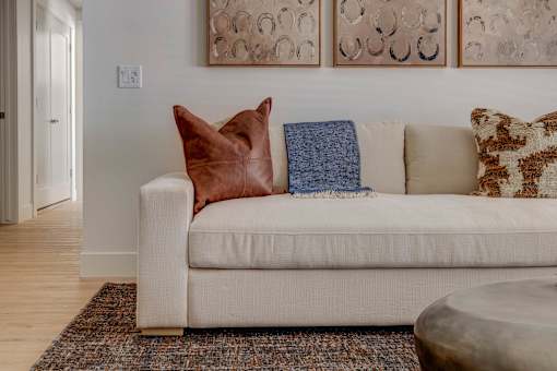 A white couch with a brown pillow and a blue blanket is in the foreground of a room with a patterned rug and a round coffee table.