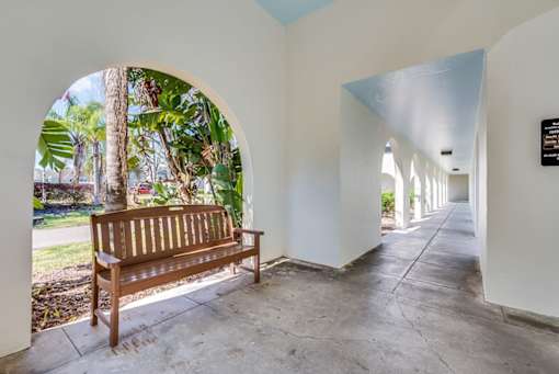 A wooden bench sits in a white hallway with a view of trees through an arched window.