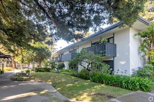 A white building with a balcony is surrounded by greenery.