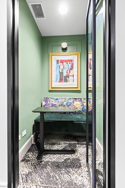 A framed picture hangs on a green wall above a table with a colorful cushion.