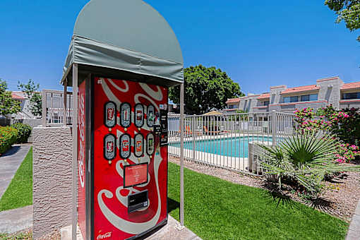 A red vending machine with a grey roof is in the foreground of a grassy area with a pool and buildings in the background.