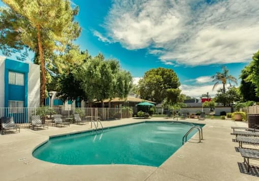 A large outdoor swimming pool surrounded by trees and a blue building at Sycamore Square Apartments, Mesa, AZ, 85202