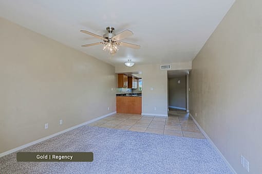 A room with a ceiling fan and a kitchen area in the background.