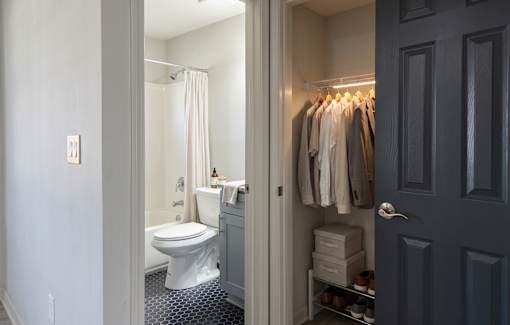 A bathroom with a toilet, a shower curtain, and a closet with clothes.