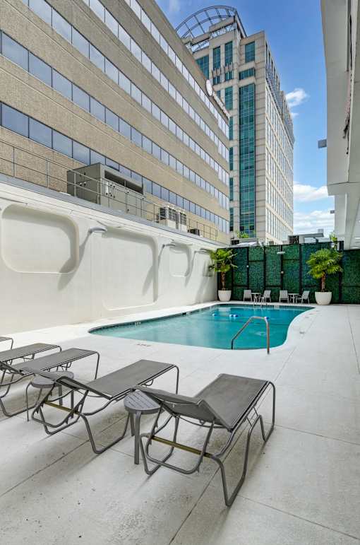 A pool with lounge chairs is surrounded by a white wall and a tall building.
