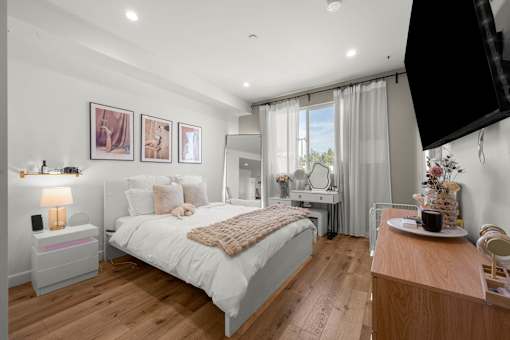 A bedroom with a large bed, a nightstand with a lamp, a dresser with a mirror, and a window with curtains.