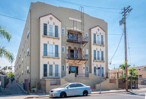 A white car is parked in front of a two-story building with a balcony.