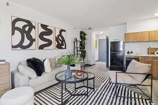 A living room with a black and white striped rug, a grey couch, a black chair, a glass coffee table, and three abstract paintings on the wall.
