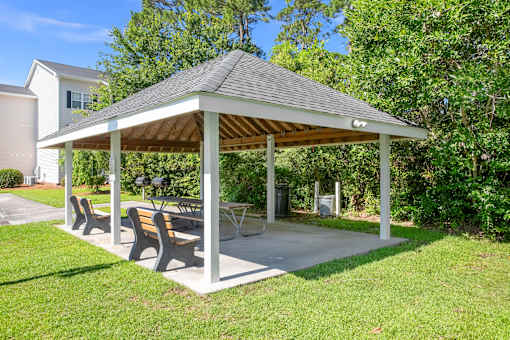 A pavilion with a roof and four benches is surrounded by greenery.