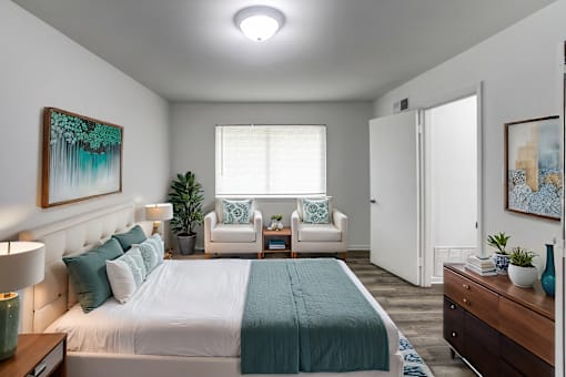 A bedroom with a bed, two chairs, a nightstand, and a painting on the wall.