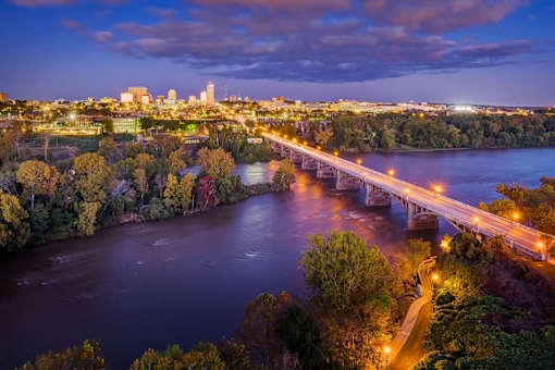 A city skyline is visible from a bridge over a river at dusk.