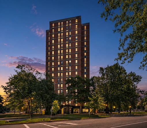 A tall building with many windows is surrounded by trees and has lights on inside.