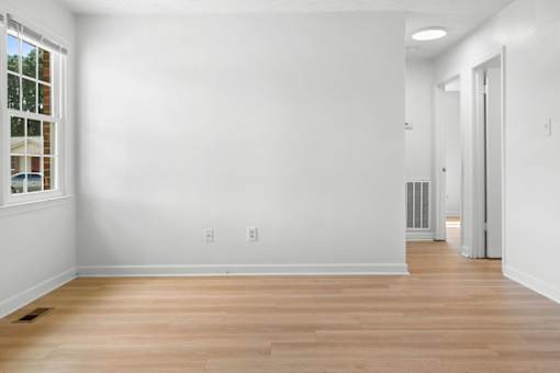 A room with white walls and wooden flooring with a window on the left side.
