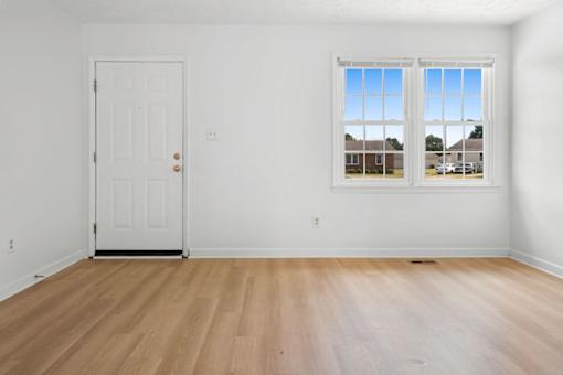 A room with a white door and a window with a view of a house across the street.