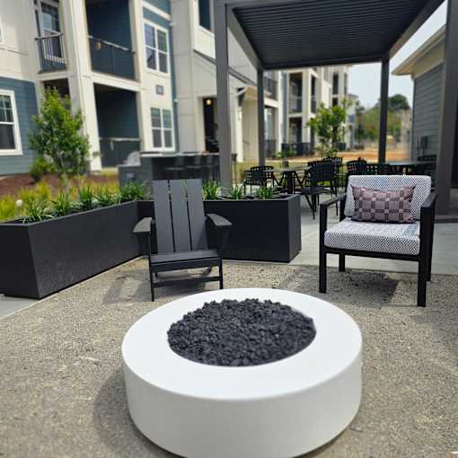 A white fire pit with black rocks in the middle sits in the middle of a patio with two chairs and a table.