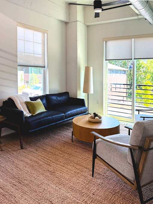 A black couch with a green pillow sits in a room with a fan and a window.
