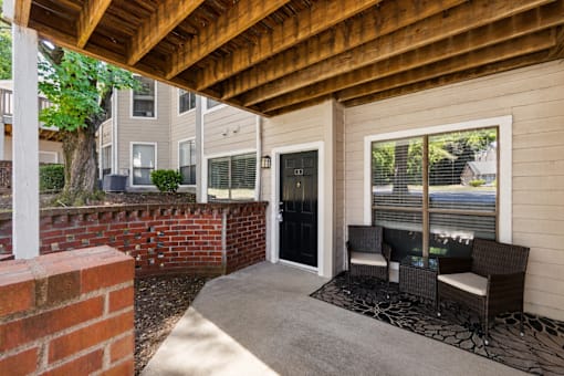 A patio with a black wicker chair and a black cushion is under a wooden roof.