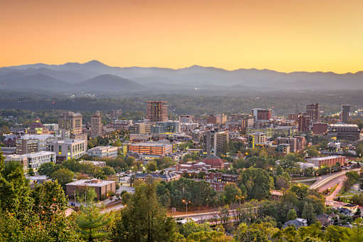A cityscape with buildings and trees in the foreground and mountains in the background.