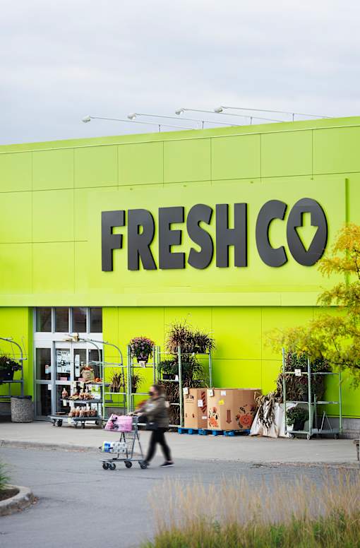 A person is pushing a shopping cart in front of a bright yellow FreshCo store.