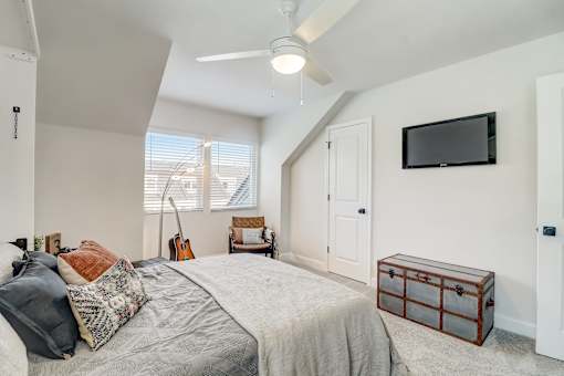 A bedroom with a bed, a chair, a guitar, a ceiling fan, and a flat-screen TV mounted on the wall.