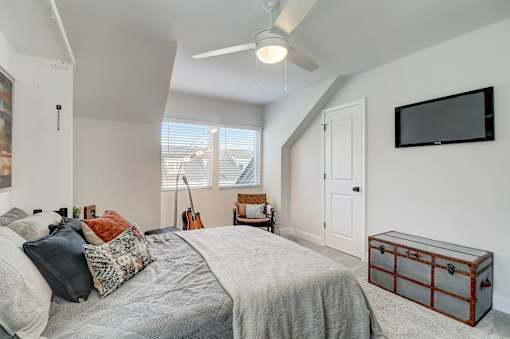 A bedroom with a bed, a guitar, a window with blinds, a ceiling fan, a wall-mounted TV, and a wooden chest.