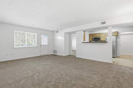A spacious living room with a carpeted floor and a kitchenette in the background.