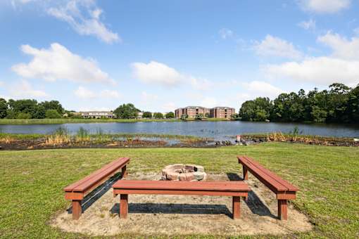 A picnic table sits in the middle of a grassy area with a pond and buildings in the background.