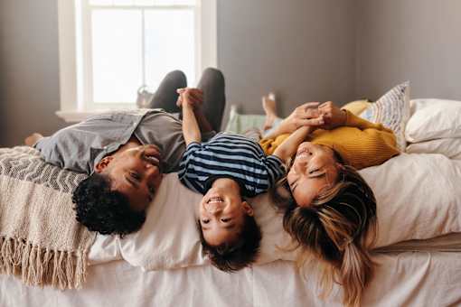 A family of three is lying on a bed, smiling and enjoying each other's company.