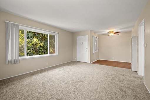 A room with a carpeted floor, a window with white curtains, and a ceiling fan.