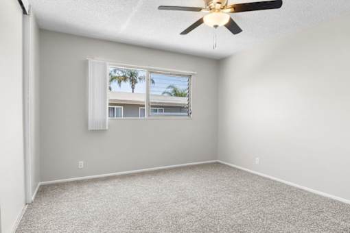 A room with a ceiling fan and a window with a view of a building and palm trees.