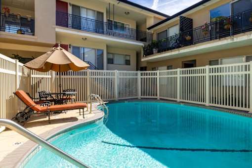 A pool with a sun lounger and umbrella in front of a white fence.
