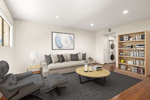 A living room with a grey couch, a grey chair, a black coffee table, a lamp, a book, a plant, a picture on the wall, a white door, and a bookshelf.