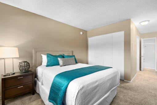 Mar Vista CA Apartments - Vista - Furnished Bedroom with Plush Carpeted Floors, Bed, Wooden Nightstand with Drawers and a Lamp, Beige Walls, and Large White Closet
