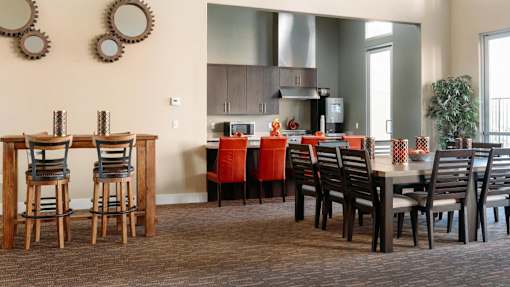 Apartments in Redlands, CA, with In-Unit Washer and Dryer - Circa 2020 - Clubhouse with High-Top Wooden Bar Table, Woven Seats, a Kitchen with Stainless Steel Appliances, and Carpeted Flooring.