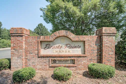 A sign for Brake Pointe Manor with a brown brick wall and bushes in front.