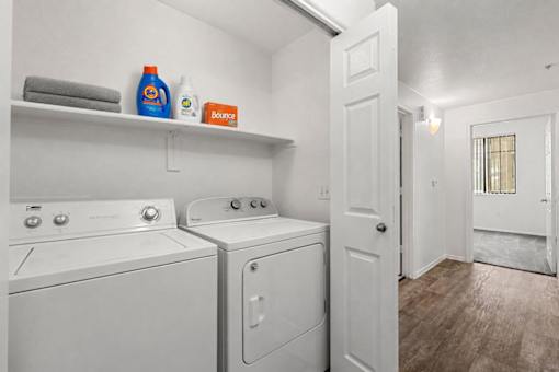 A laundry room with a white washer and dryer, a shelf with towels and cleaning products, and a doorway leading to another room.