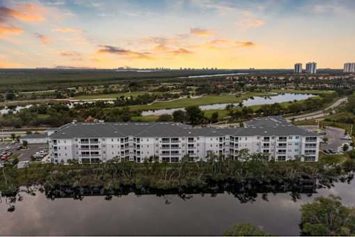 A large white building is situated on the edge of a body of water with a parking lot in front.