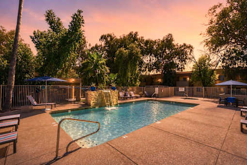 A pool surrounded by trees and chairs with a sunset in the background.