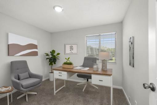 A room with a grey chair, a desk with a plant on it, and a painting on the wall.