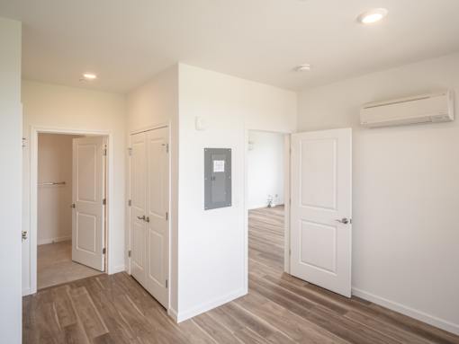 A room with white walls and wooden floors has a white door and a white cabinet with a mirror above it.