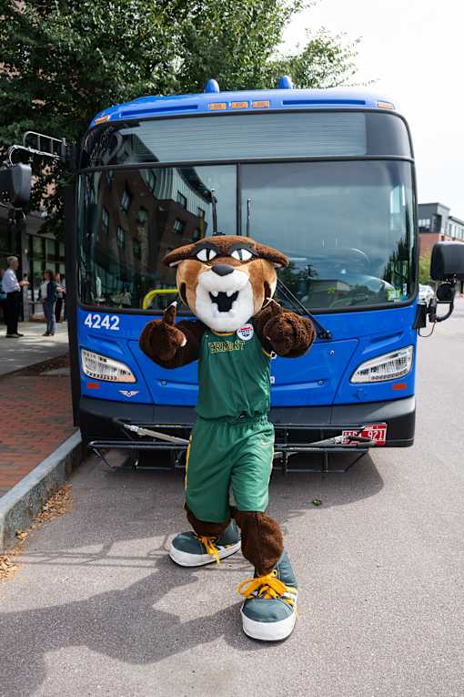 A person in a mascot costume stands in front of a blue bus.